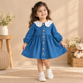 A young girl smiling and wearing a blue denim dress with a white lace Peter Pan collar and gold buttons, standing in a warm, decorated room with a teddy bear and wooden furniture.