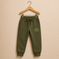 Olive green jogger sweatpants with a drawstring waist and 'Stay Cool' text graphic on the left leg, hanging on a wooden hanger against a solid beige background.