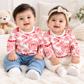 Two babies sitting side by side on a white fluffy rug, wearing matching white long-sleeve tops with red graphic prints and jeans or cream pants. The baby on the left wears blue jeans and white socks, while the baby on the right wears cream pants, white socks, and a cream bow headband.