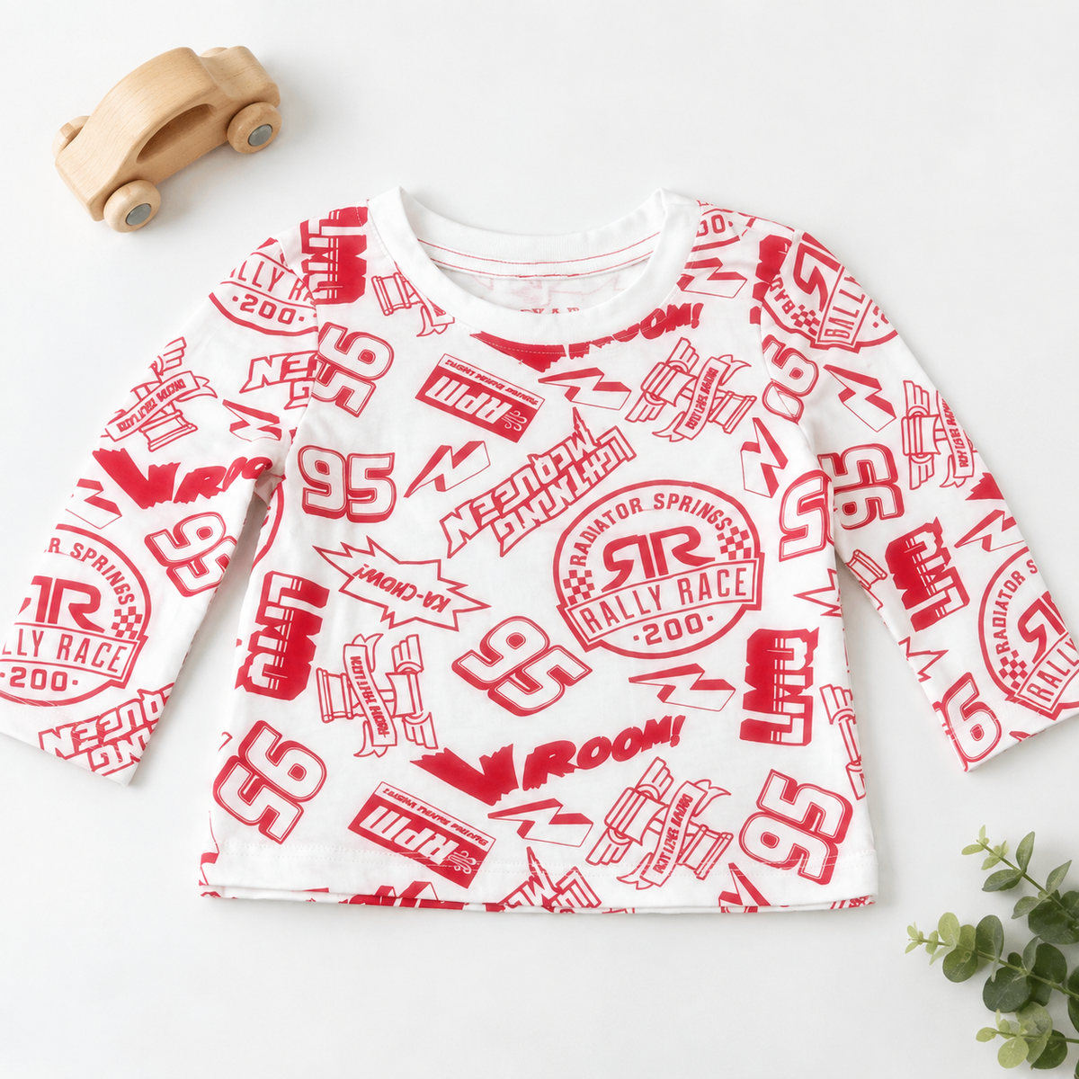 A baby long-sleeve t-shirt with a red and white racing graphic print, styled in a flat-lay with a wooden toy car and green leaves on a white background.